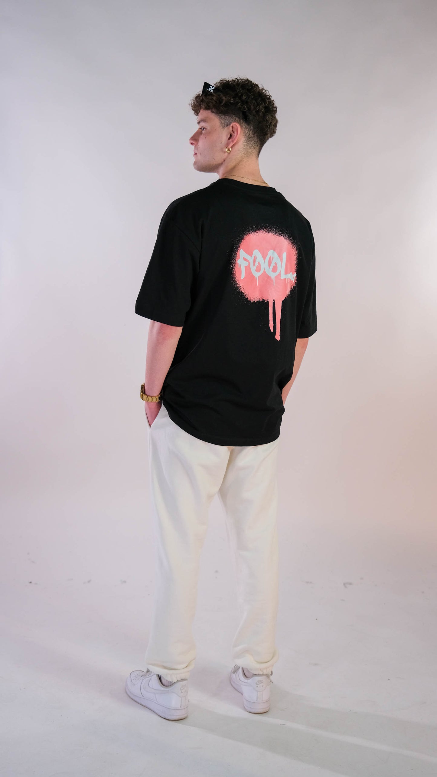 Achterkant mannelijk model met zwart t-shirt met 'Graffiti FØØL' logo op de rug. Ook draagt hij de witte 'FØØL' jogger.
