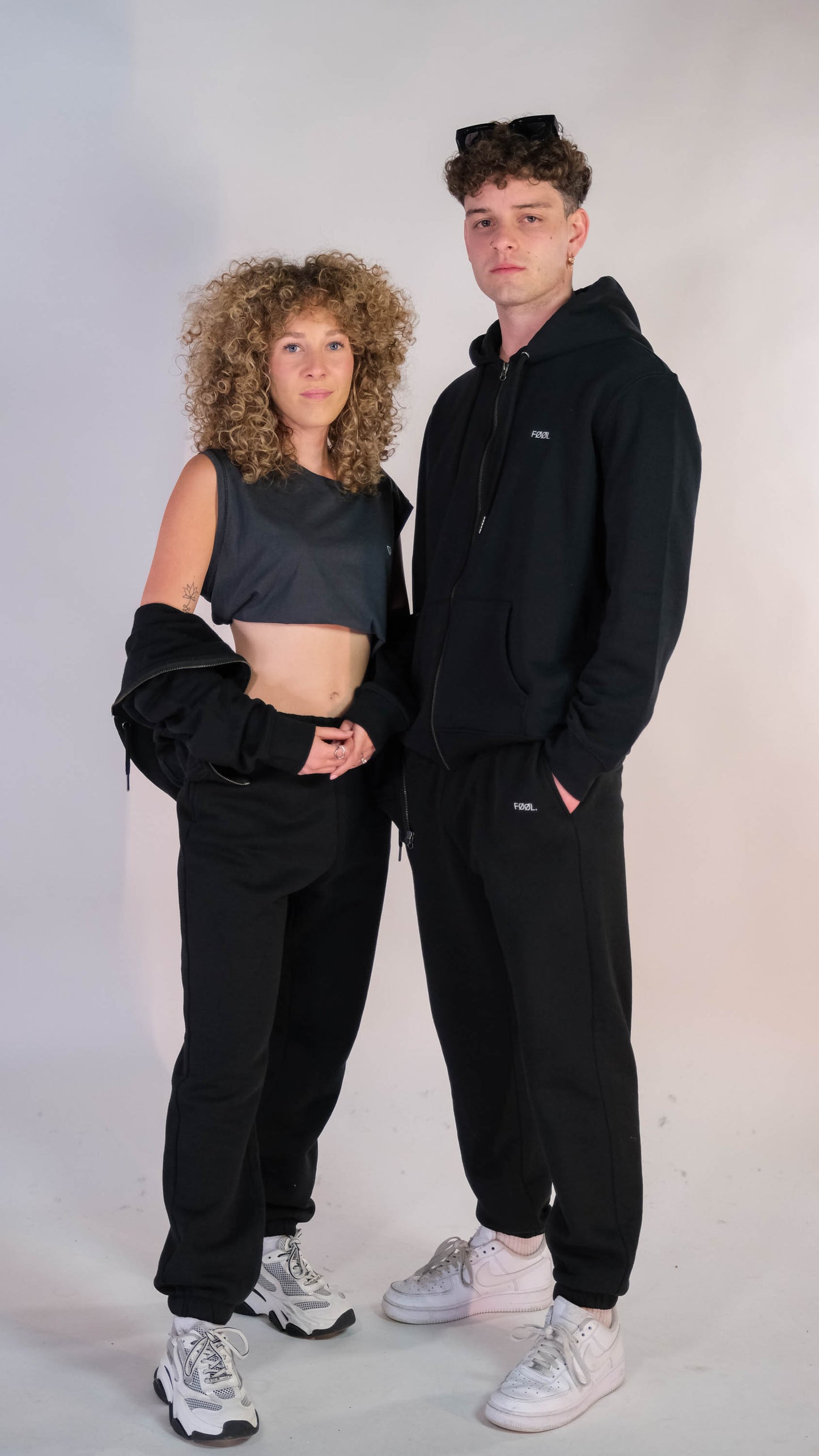 2 modellen. Vrouw draag zwart vest los over haar armen, man draagt zwart 'FØØL' vest dicht. Beide modellen dragen een zwarte 'FØØL' jogger.