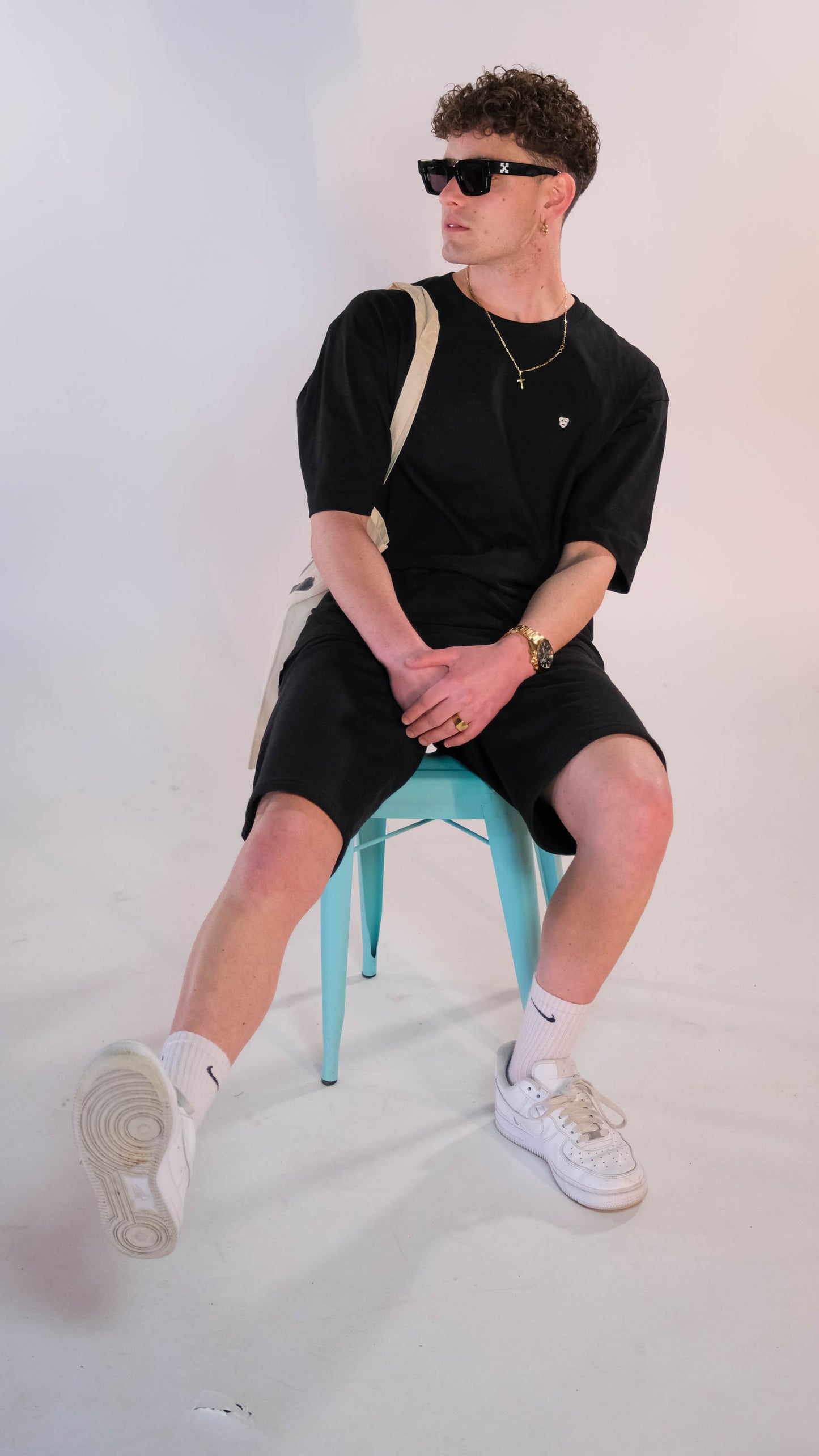 Mannelijk model met zwart t-shirt, wit 'BEAR' logo op de borst, zitten op een blauwe kruk. Hij draagt de zwarte 'FØØL' shorts.
