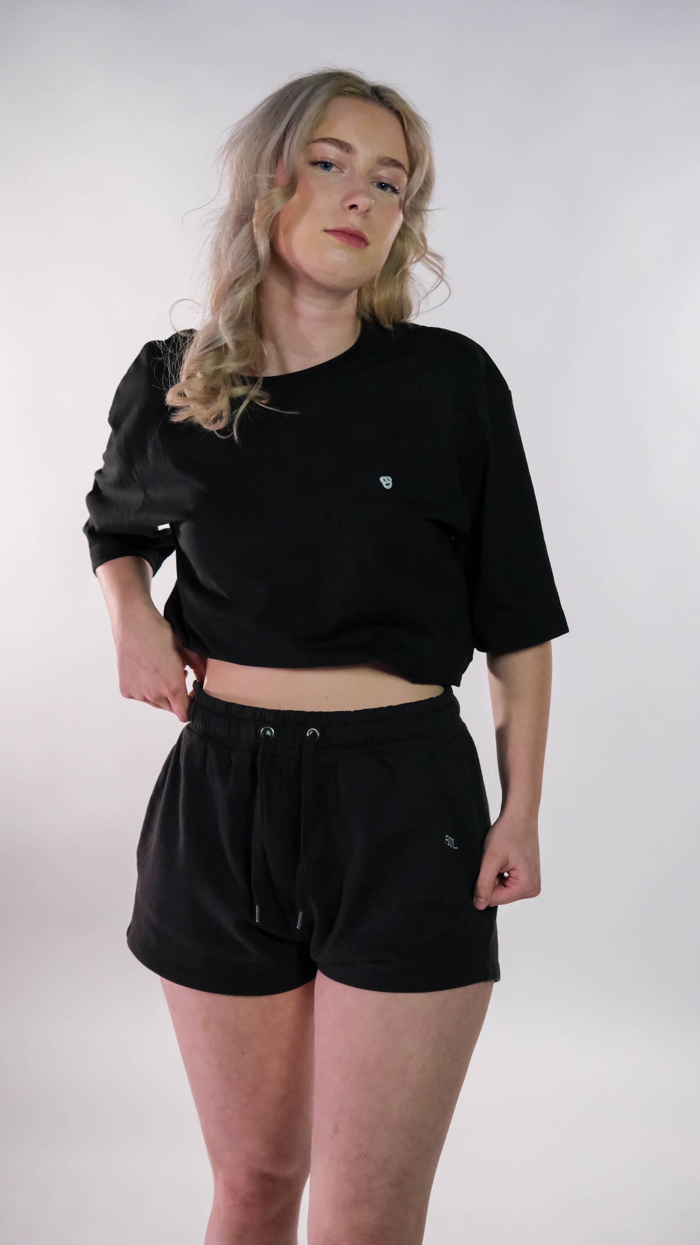 Vrouwelijk model met zwart t-shirt, wit 'BEAR' logo op de borst, waarbij zij het t-shirt wat naar binnen heeft opgekruld. Zij draagt de zwarte 'FØØL' shorts.