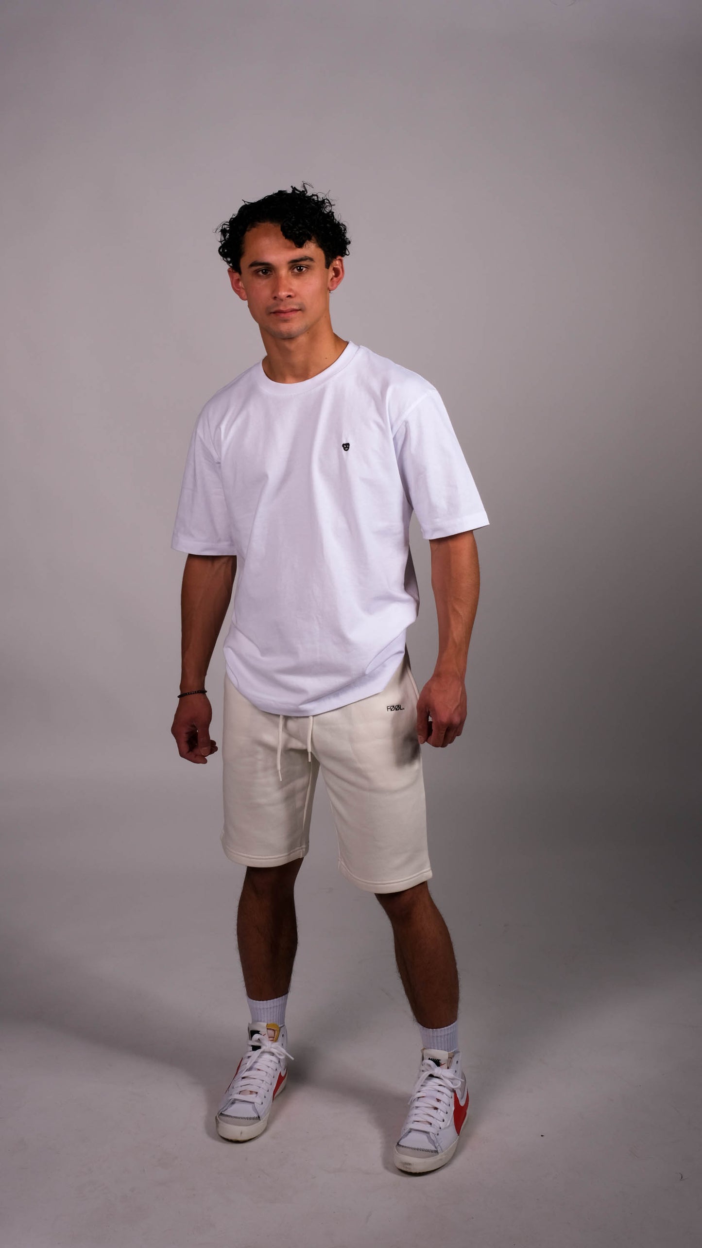 Mannelijk model met wit t-shirt met zwart 'BEAR' logo op de borst. Hij draagt de witte 'FØØL' shorts.