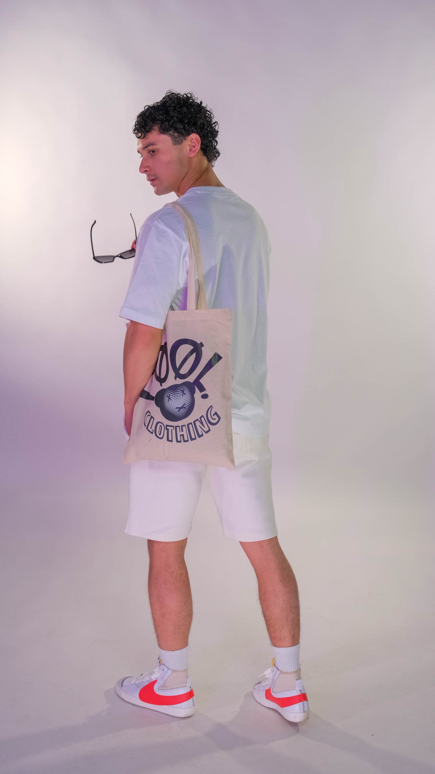 Achterkant mannelijk model met wit t-shirt. Model draagt een witte 'FØØL' shopper over de schouder en heeft een zonnebril in zijn hand. Hij draagt de witte 'FØØL' shorts.