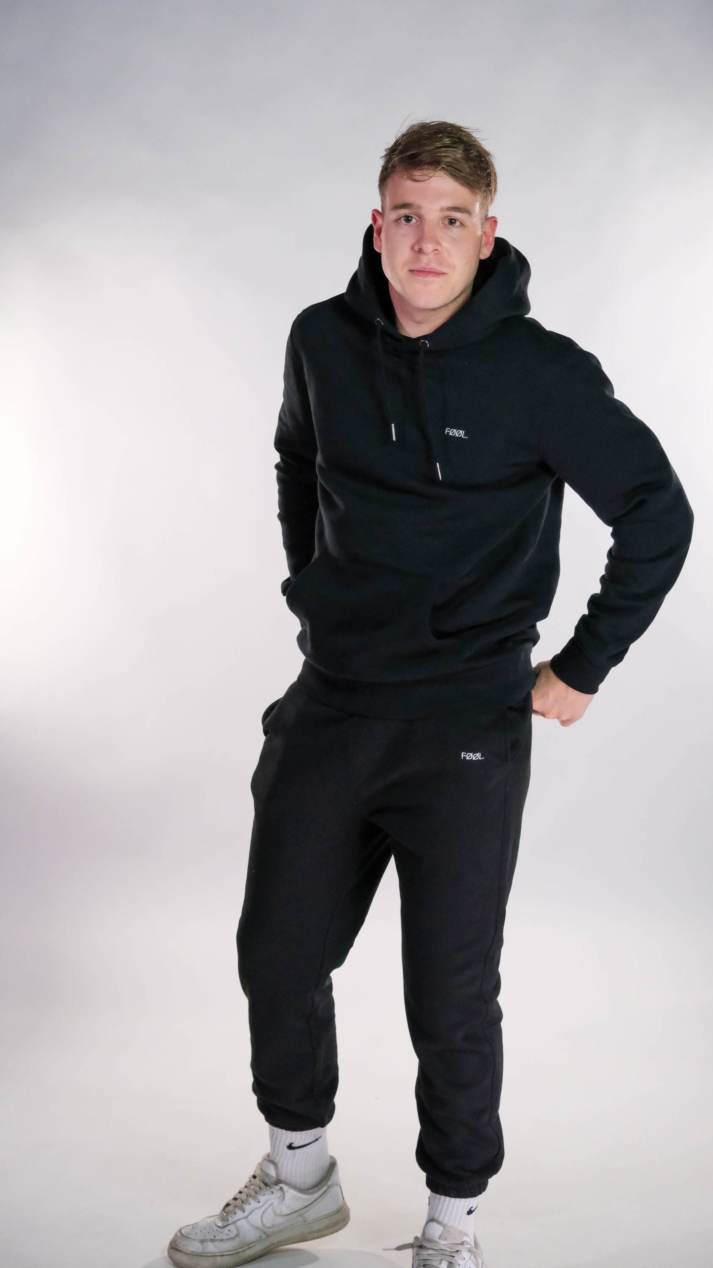 Jongen draagt een zwarte hoodie en zwarte jogger, beide voorzien van een wit 'FØØL' logo.