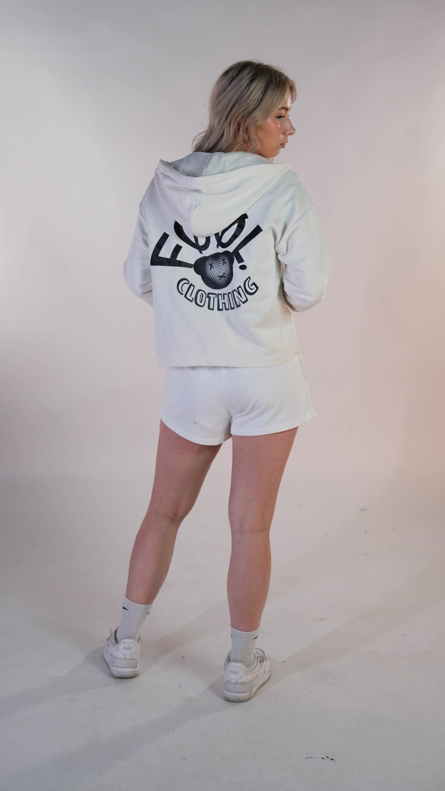 Vrouwelijk model die met het oversized cropped vest, in de kleur ivory, zwart ' FØØL CLOTHING' draagt. Zij staat met haar rug naar de camera.