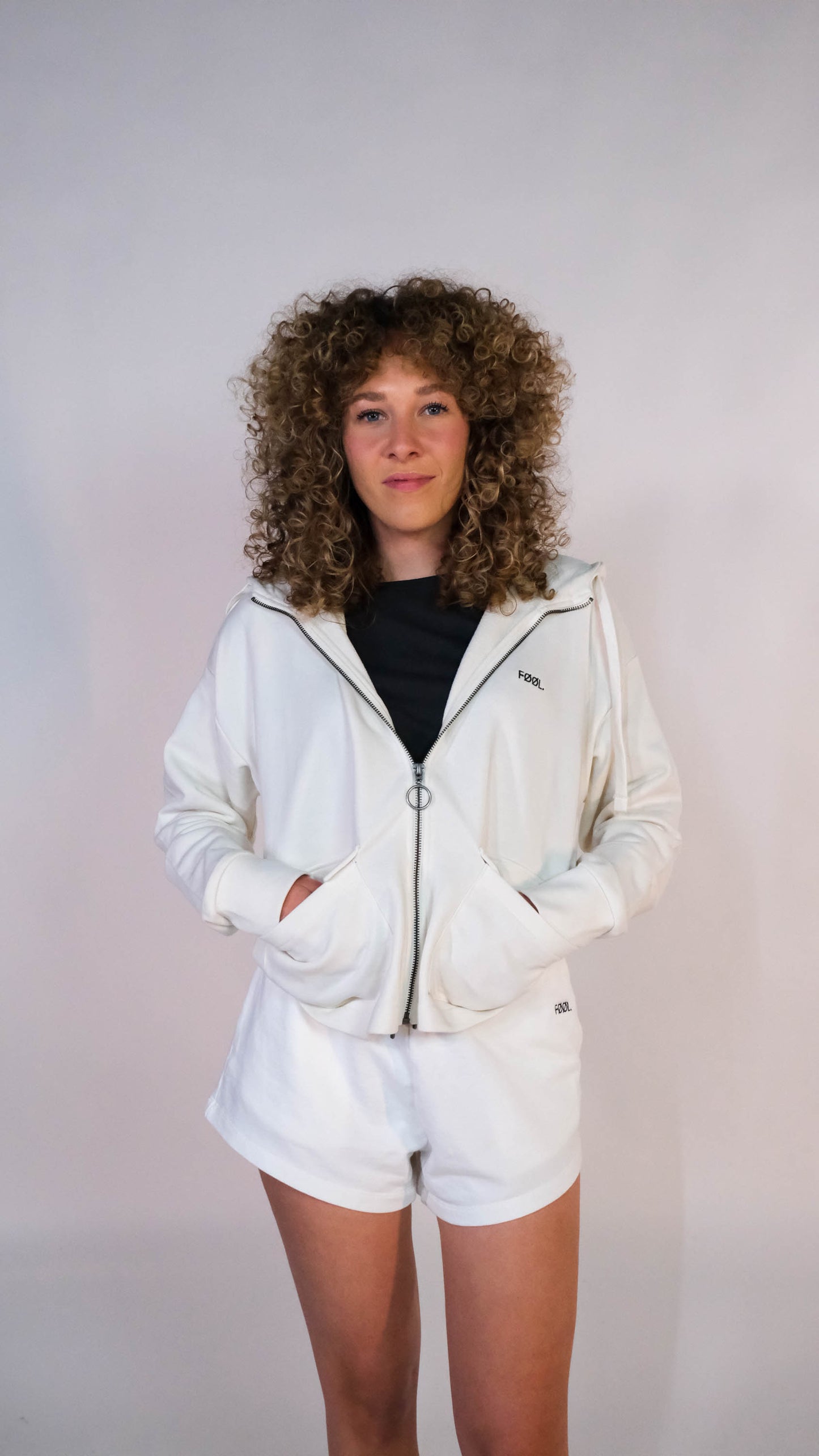 Vrouwelijk model met het oversized cropped vest, half dicht geristst naar de camera. Zij draagt het vest in de kleur ivory met het zwarte 'FØØL' logo op de borst en de ivory shorts.