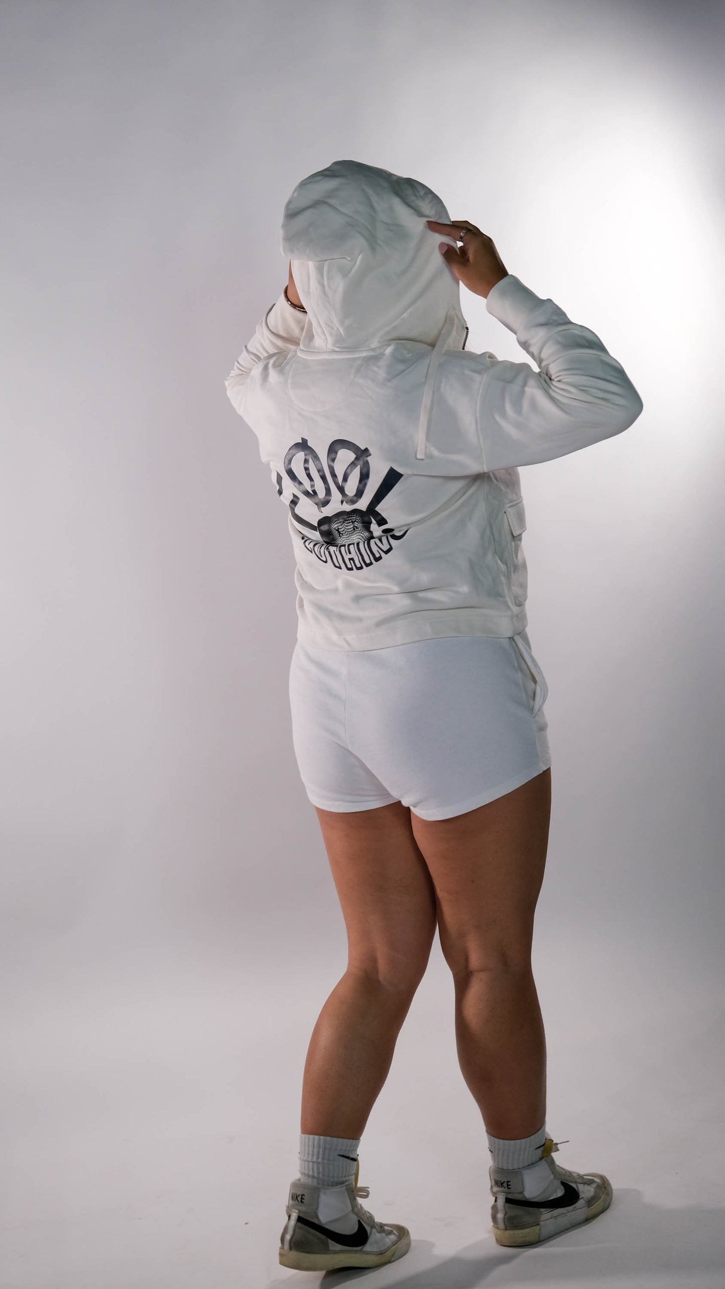 Een model poseert met de rug naar de camera en draagt het oversized cropped vest in de kleur ivory met het "Fool Clothing" logo op de achterkant, gecombineerd met de witte 'FØØL' short. Het model houdt de capuchon vast en draagt hoge sokken en versleten Nike sneakers, wat de urban streetwear look compleet maakt.