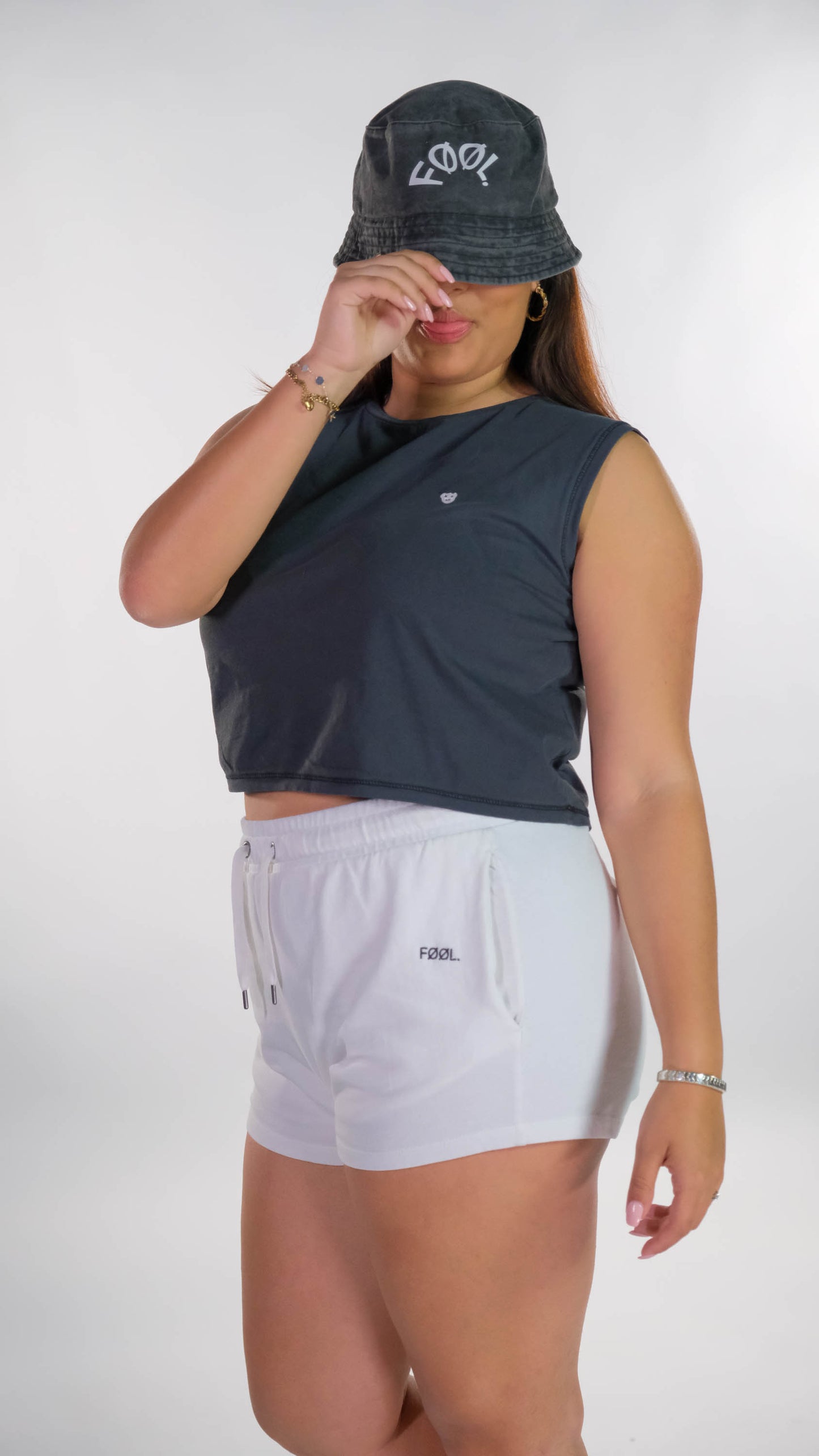 Vrouwelijk model draagt de verwassen zwarte cropped top met de witte 'BEAR' logo op de borst en een witte short met het zwarte 'FØØL' logo aan de zijkant. Ook draagt zij de verwassen zwarte bucket hat met het witte 'FØØL' logo die zij lichtelijk naar beneden trekt.
