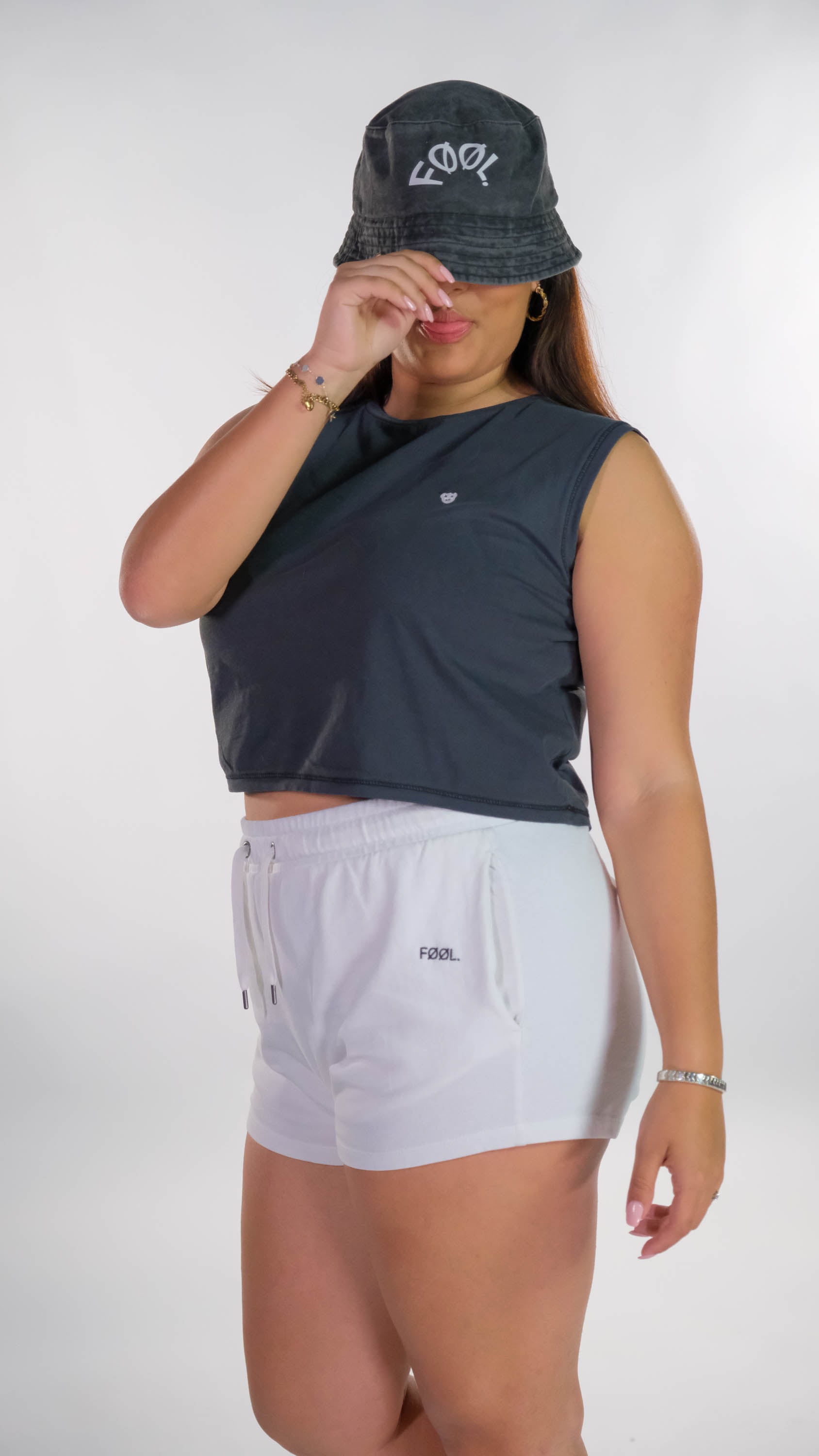 Vrouwelijk model draagt de verwassen zwarte cropped top met de witte 'BEAR' logo op de borst en een witte short met het zwarte 'FØØL' logo aan de zijkant. Ook draagt zij de verwassen zwarte bucket hat met het witte 'FØØL' logo die zij lichtelijk naar beneden trekt.