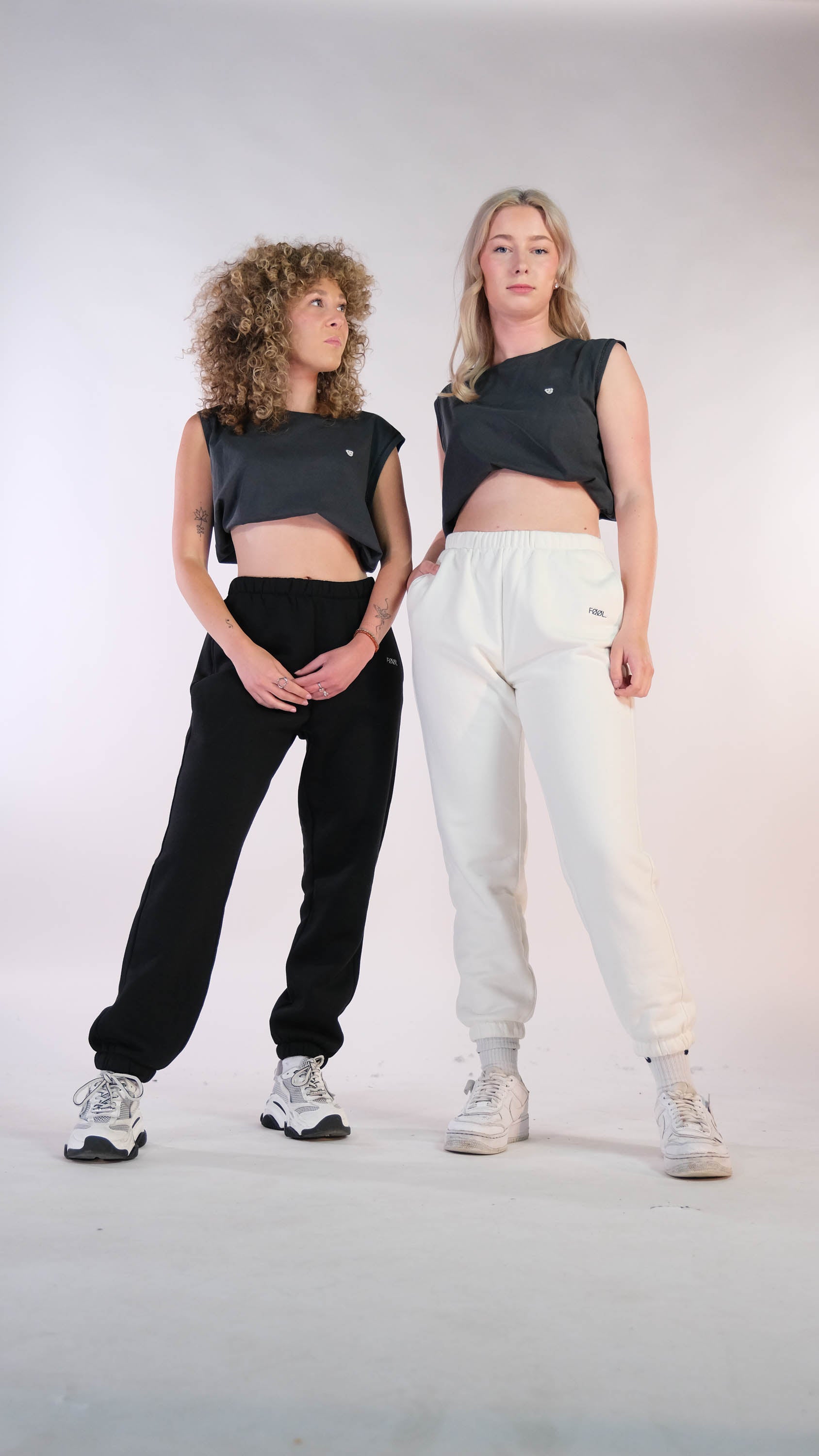 2 vrouwelijke modellen die beide de verwassen zwarte cropped top met wit 'BEAR' logo dragen. Model links draagt daar de zwarte 'FØØL' jogger bij. Model rechts draagt daar de ivory 'FØØL' jogger bij.