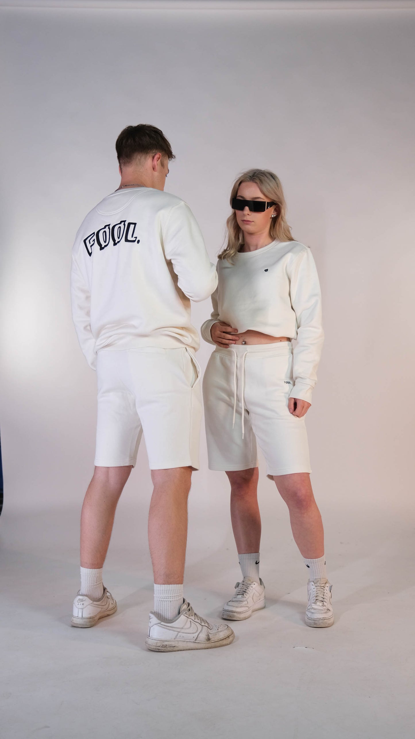 2 modellen. Het vrouwelijke model met de ivory 'FØØL' sweater met het zwarte 'BEAR' logo op de borst en draagt daarbij de ivory 'FØØL' short. Het mannelijk model draagt hetzelfde maar staat met haar rug naar de camera toe.