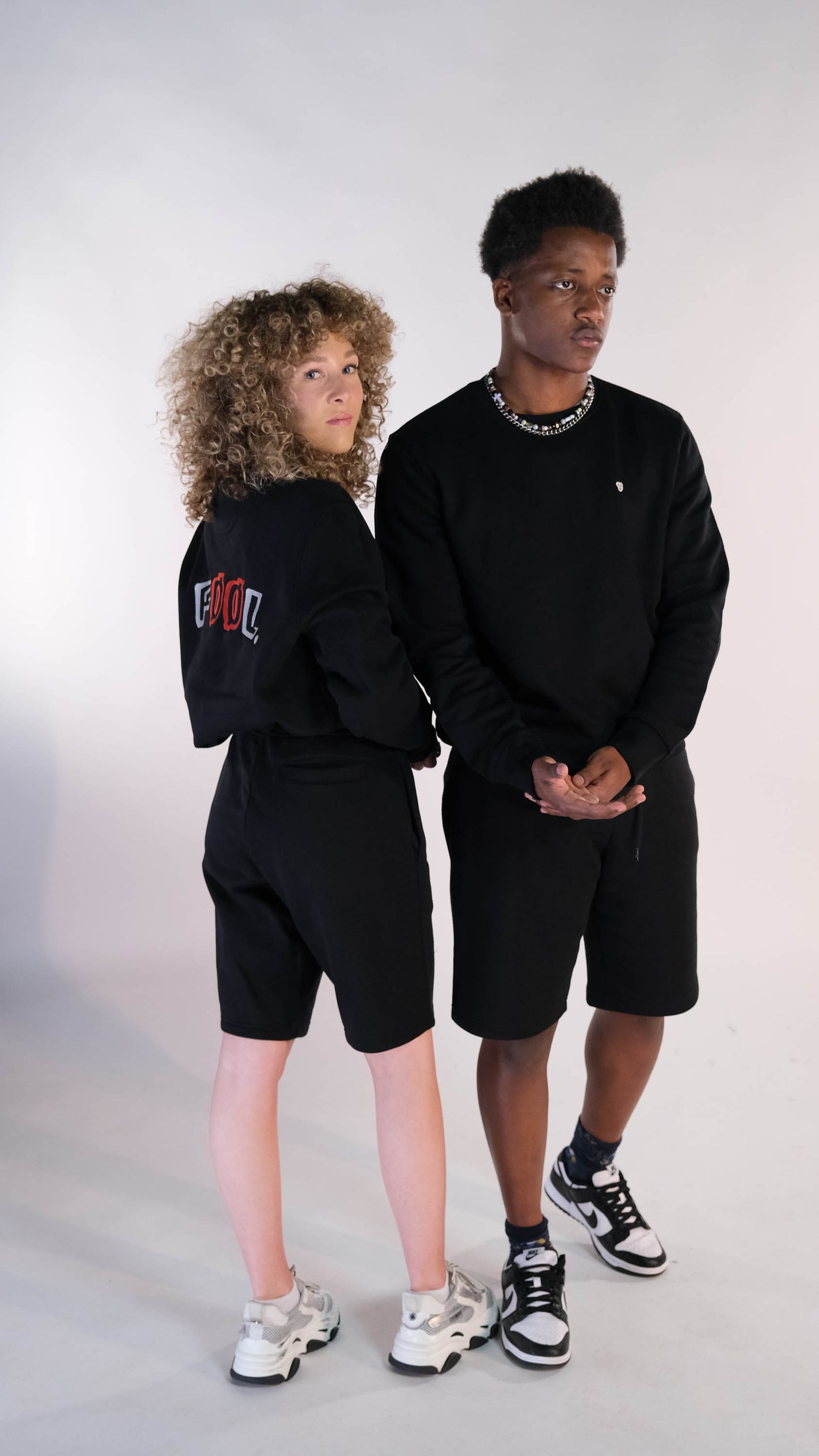 2 modellen. Een mannelijk model met de zwarte 'FØØL' sweater met het witte 'BEAR' logo op de borst en draagt daarbij de zwarte 'FØØL' short. Het vrouwelijke model draagt hetzelfde maar staat met haar rug naar de camera toe.