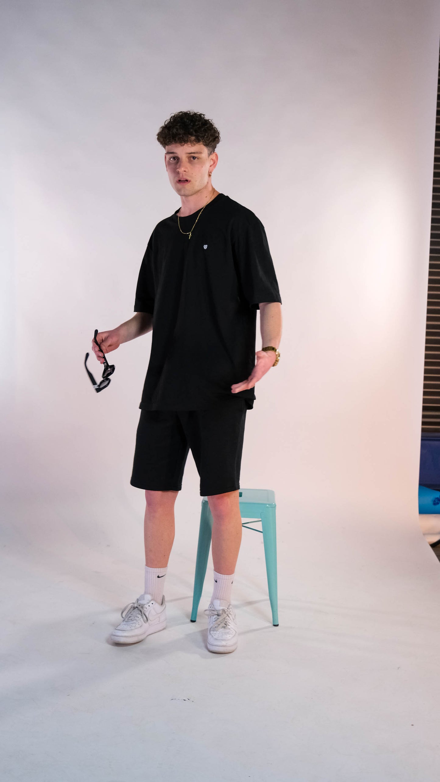 Mannelijk model met een zwart t-shirt met het witte 'BEAR' logo op de borst. Hij draagt de zwarte shorts. Hij heeft een zonnebril in zijn rechterhand.