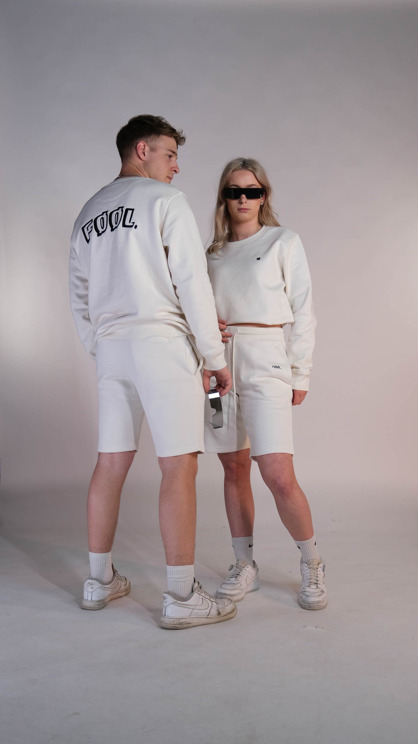 2 modellen. Het mannelijk model staat met zijn rug naar de camera. Hij draagt de ivory sweater met het zwarte 'FØØL' logo op de rug. Ook draagt hij de ivory shorts. het vrouwelijke model draagt precies hetzelfde alleen staat zij met haar gezicht naar de camera gericht.