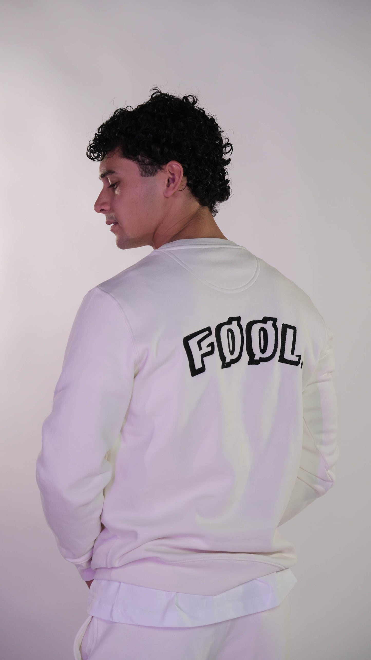 Mannelijk model die met zijn rug naar de camera toe gedraaid staat. Hij draagt de ivory sweater met het zwarte 'FØØL' logo op de rug.