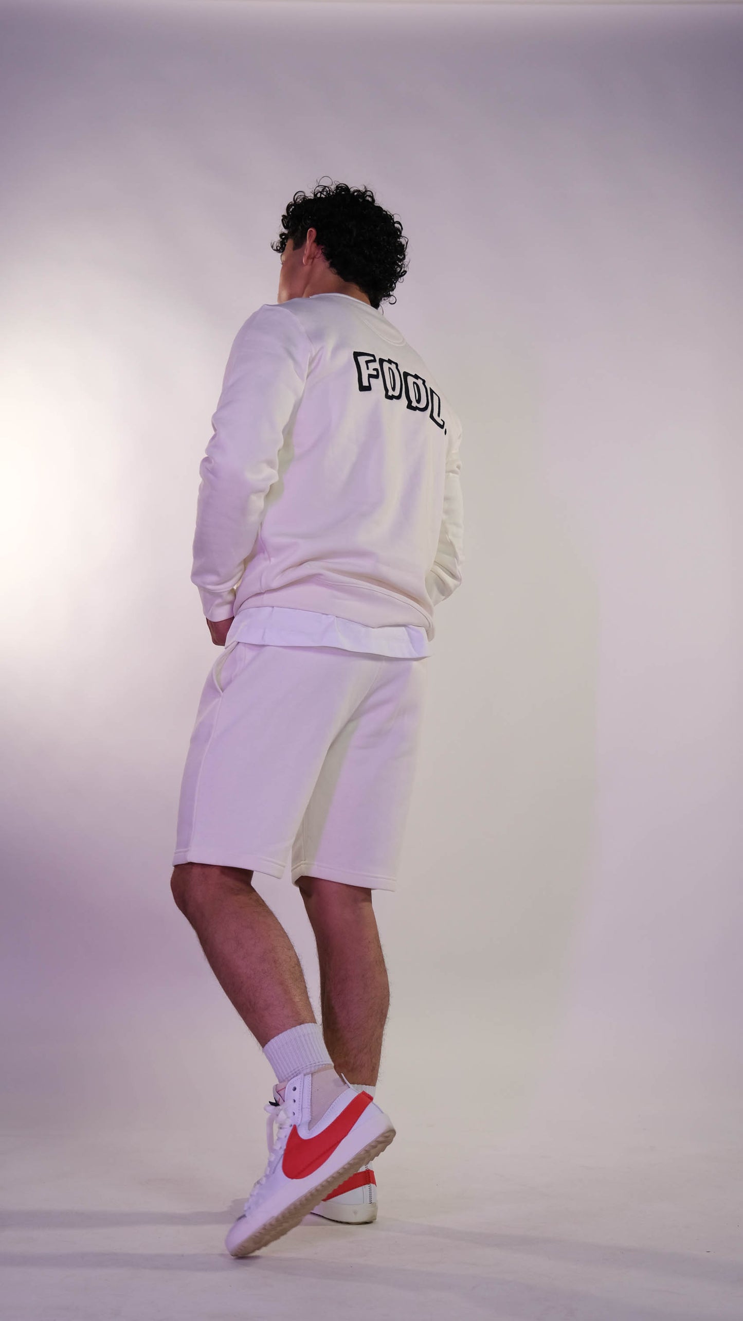 Mannelijk model die met zijn rug naar de camera toe gedraaid staat. Hij draagt de ivory sweater met het zwarte 'FØØL' logo op de rug.