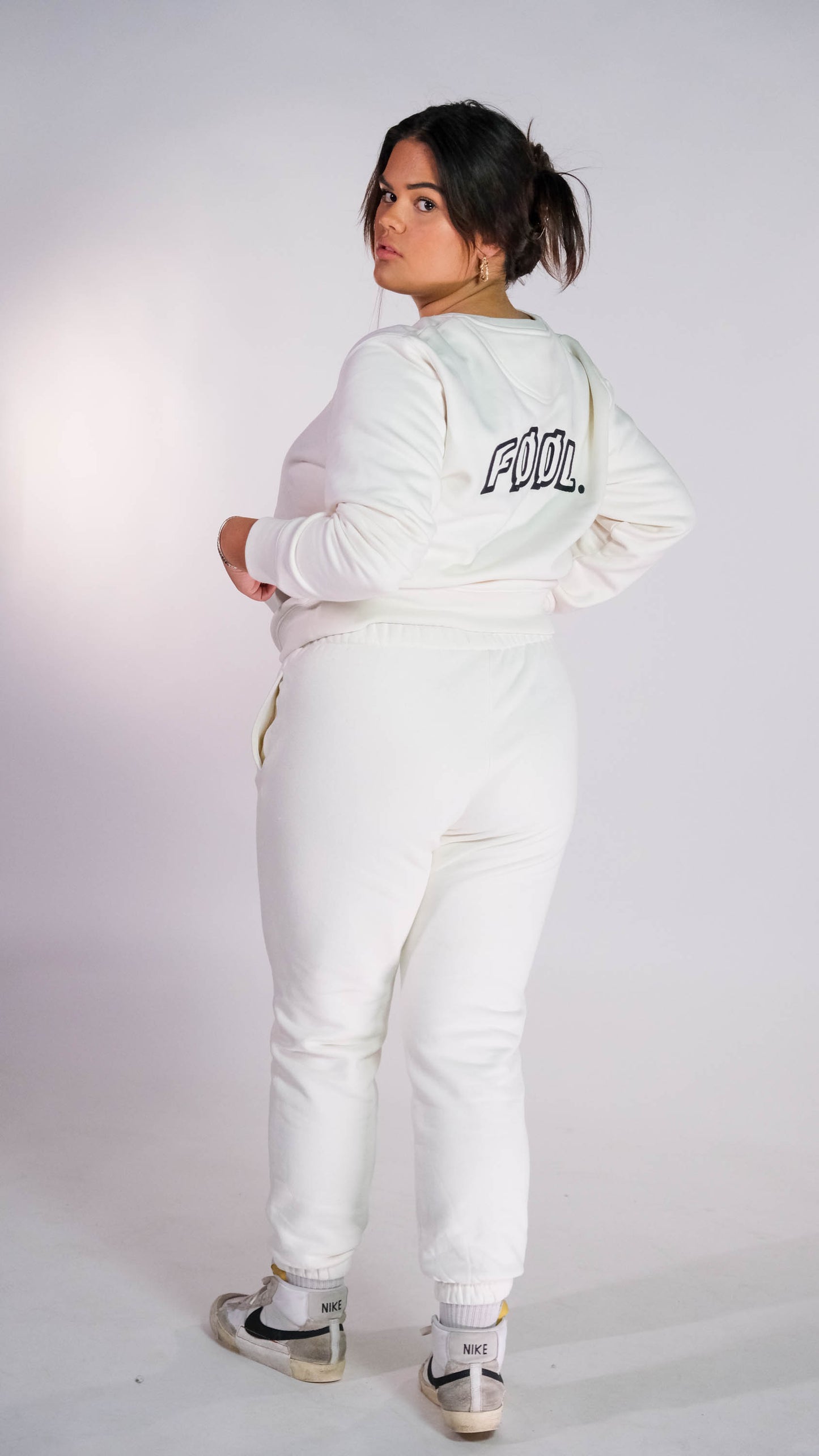 Vrouwelijk model die met haar rug naar de camera toe gedraaid staat. zij draagt de ivory sweater met het zwarte 'FØØL' logo op de rug. Ook draagt zij de ivory jogger.