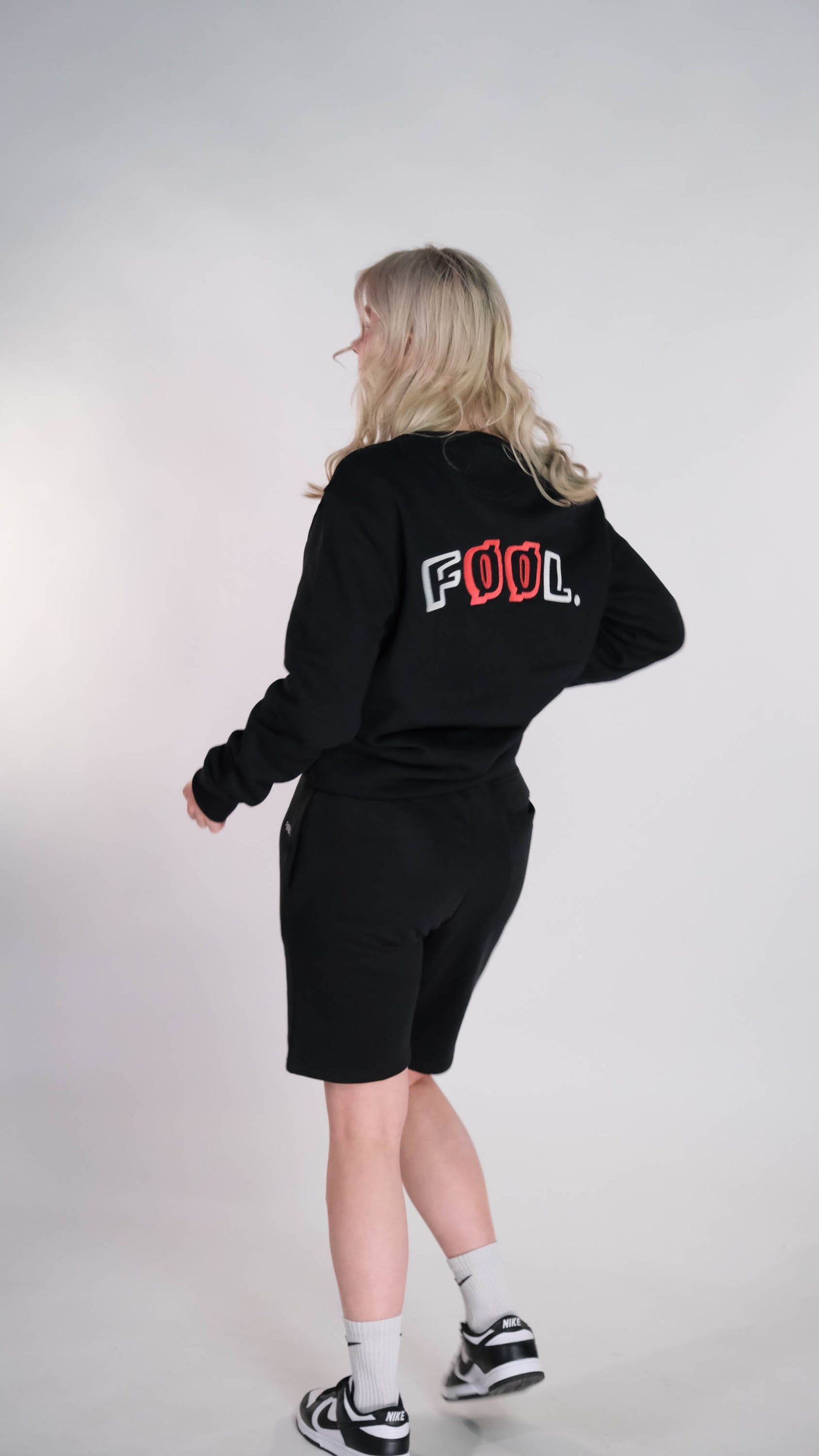 Vrouwelijk model met de zwarte sweater met het 'FØØL' logo op haar rug. Ook draagt zij de zwarte 'FØØL' short.
