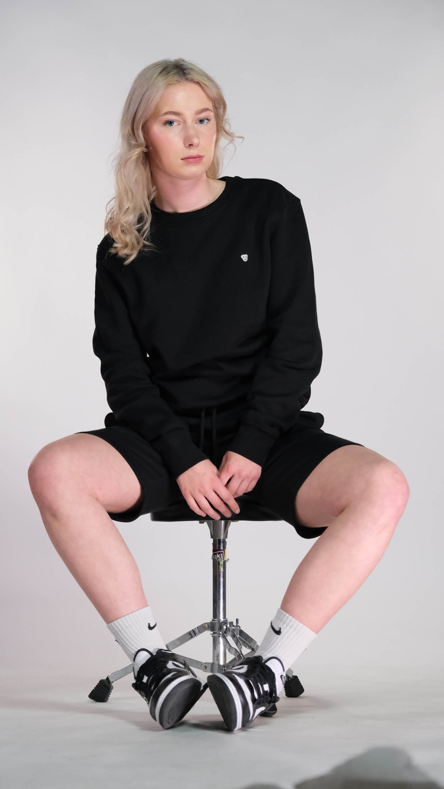 Vrouwelijk model, zitten op een kruk. Zij draagt de zwarte sweater met de witte 'BEAR' logo op de borst. Ook draagt zij de zwarte 'FØØL' short.