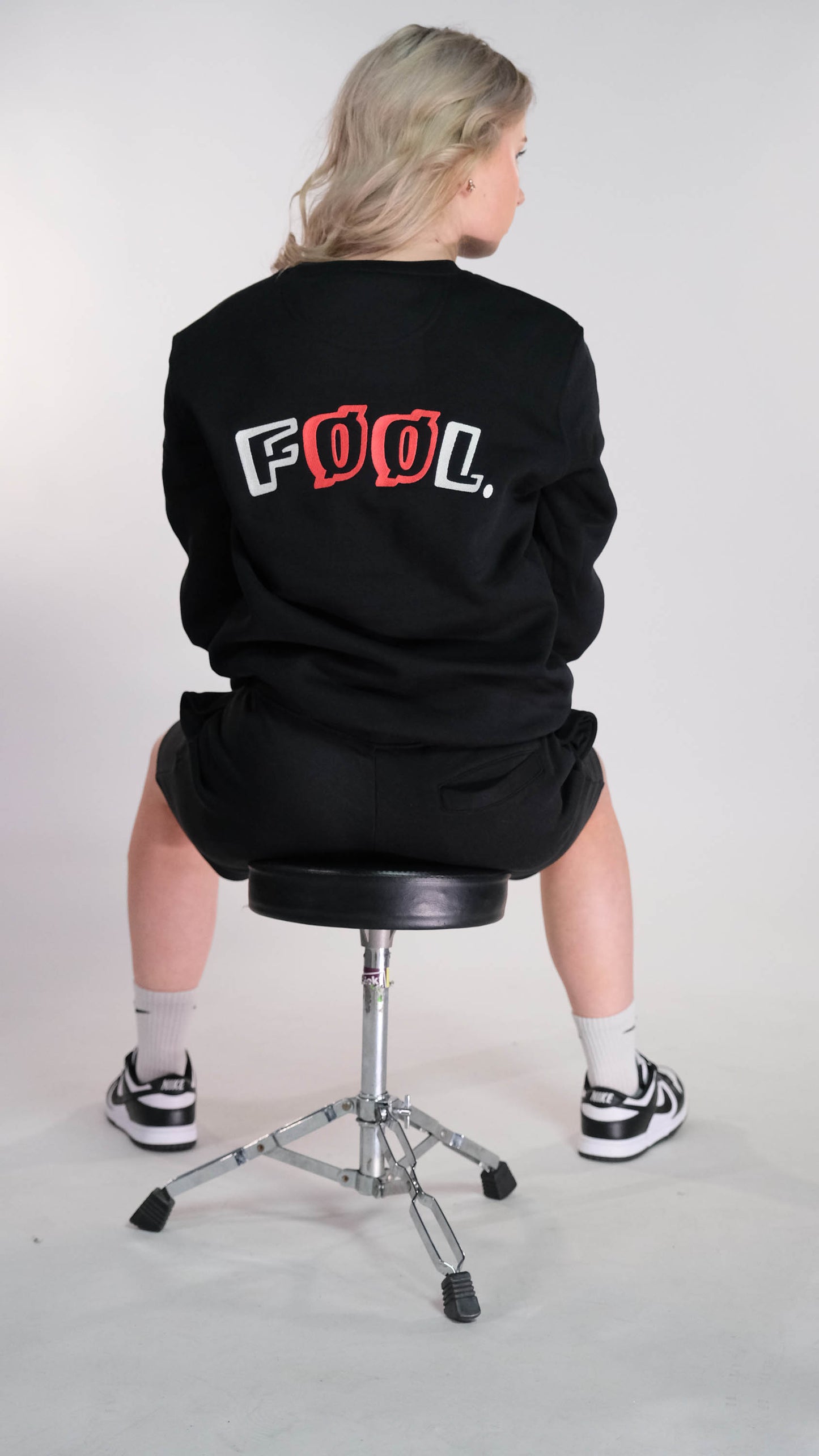 Vrouwelijk model, zittend op een kruk met haar rug naar de camera gedraaid. Zij draagt de zwarte sweater met het ivery/rode logo op de rug. Ook draagt ze de zwarte 'FØØL' short.
