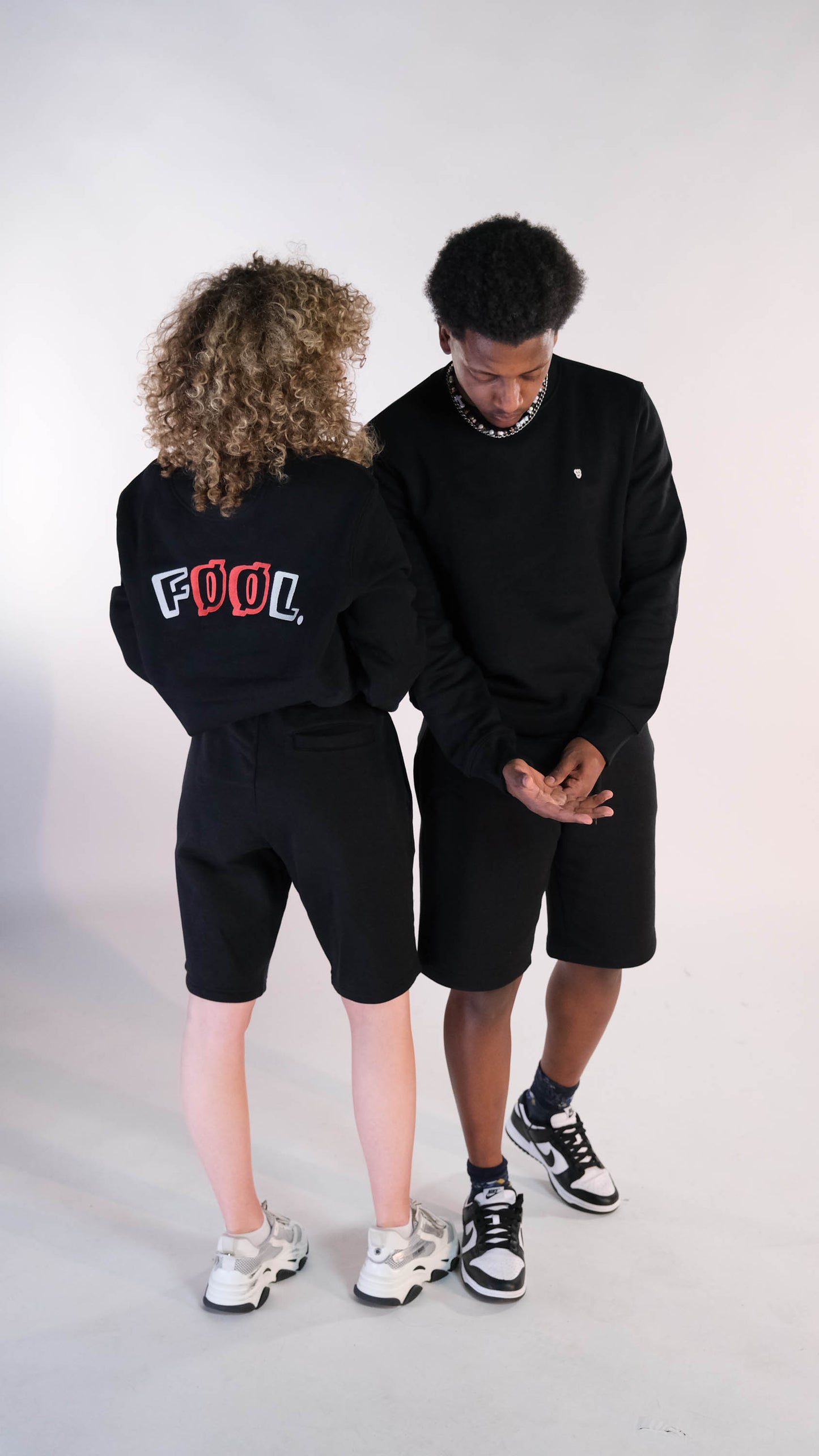 2 modellen, een man en een vrouw. De vrouw staat met haar rug naar de camera. Zij draagt de zwarte sweater met het ivory/rode logo op de rug. Ook draagt zij de zwarte 'FØØL' short. Het mannelijk model staat met zijn gezicht naar de camera toe en draagt hetzelfde als het vrouwelijk model.