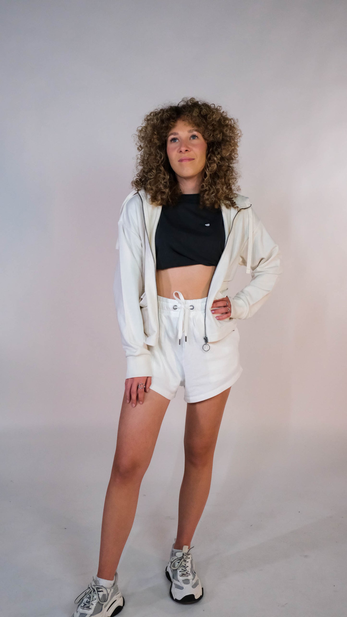 Vrouwelijk model met het oversized cropped vest, half dicht geristst naar de camera. Zij draagt het vest in de kleur ivory met het zwarte 'FØØL' logo op de borst en de ivory shorts.