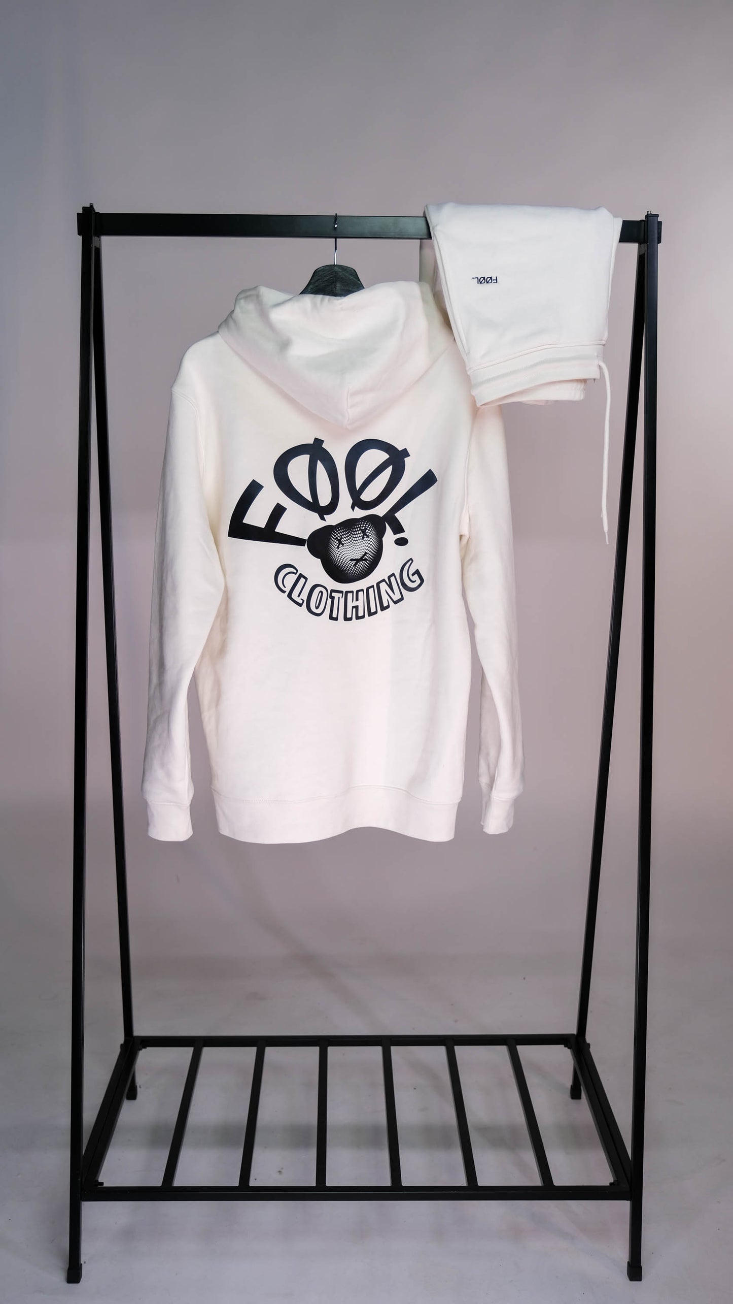 Een zwart, ijzeren kledingrek met een ivory hoodie met zwart 'FØØL CLOTHING' logo op de rug, hangend aan een kleerhanger, Daarnaast een ivory jogger met zwart 'FØØL' logo die over het ijzeren kleding rek is gehangen.