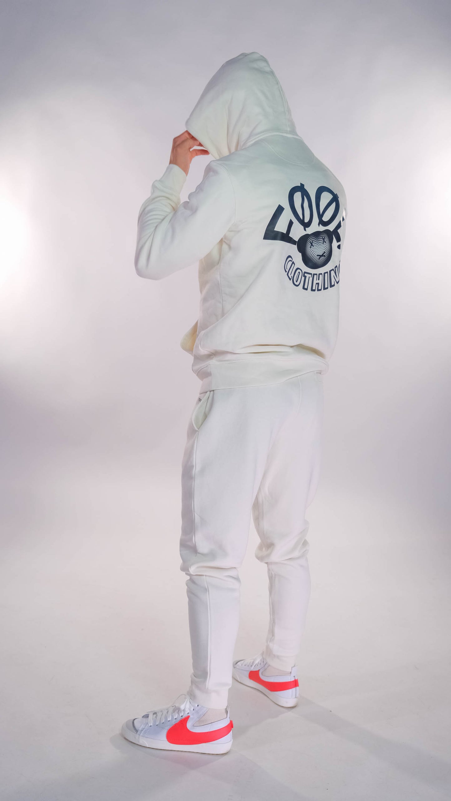 Mannelijk model die met zijn rug naar de camera gedraaid staat. Hij draagt de ivory hoodie met het zwarte 'FØØL CLOTHING' logo op de rug. Ook draagt hij de ivory jogger. Hij heeft de capuchon op.