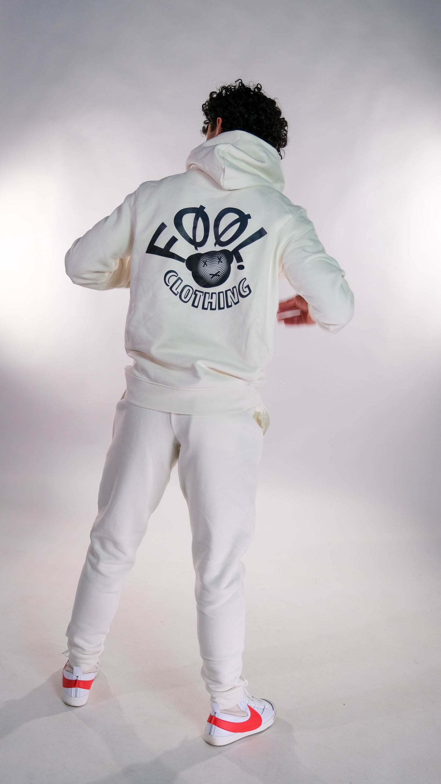 Mannelijk model die met zijn rug naar de camera gedraaid staat. Hij draagt de ivory hoodie met het zwarte 'FØØL CLOTHING' logo op de rug. Ook draagt hij de ivory jogger.