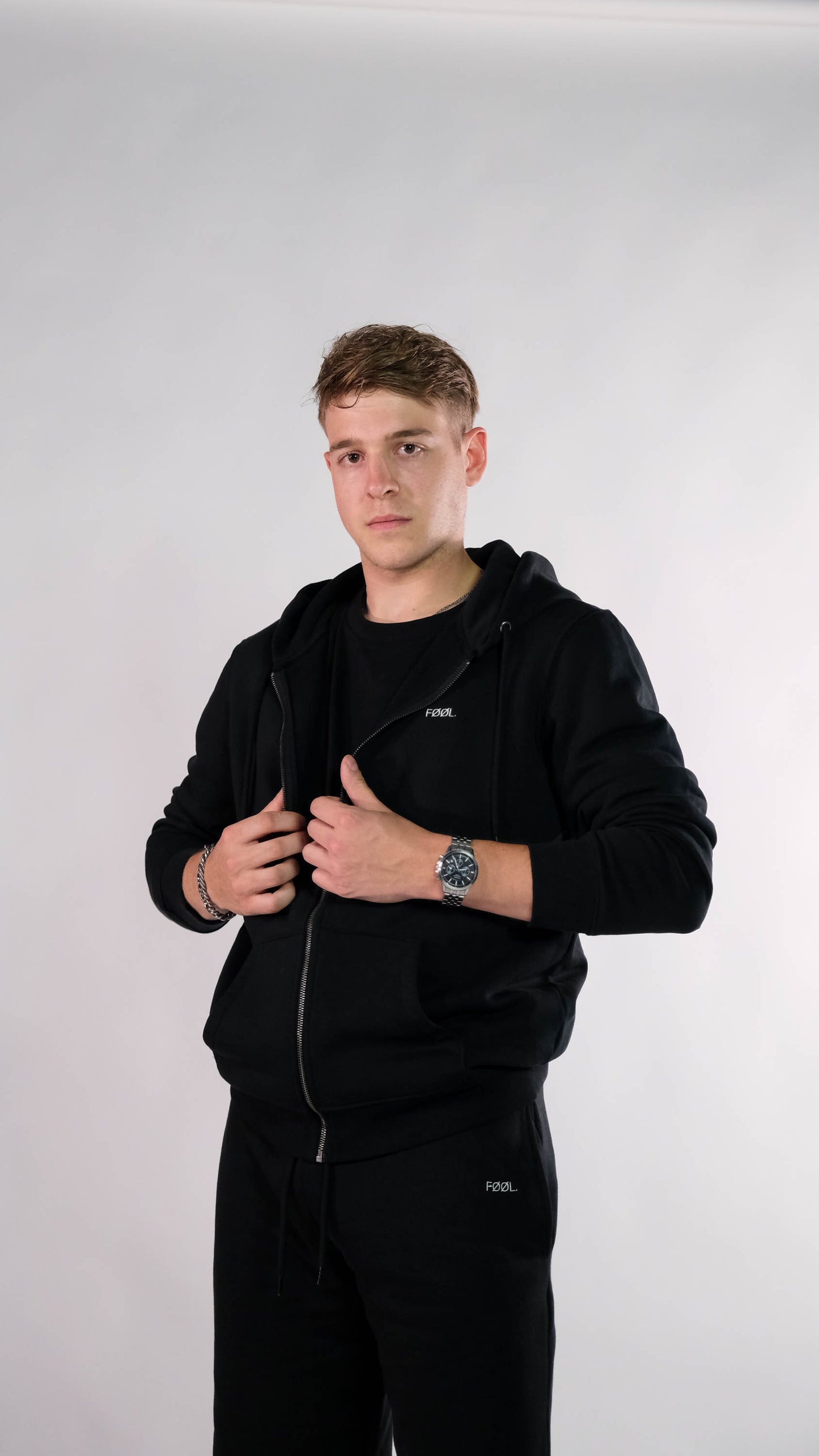 Mannelijk model met zwart vest met wit 'FØØL' logo op de borst. Hij draagt ook de zwarte 'FØØL' jogger.